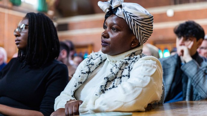 A Black woman audience member in a headscarf is seated and listening attentively