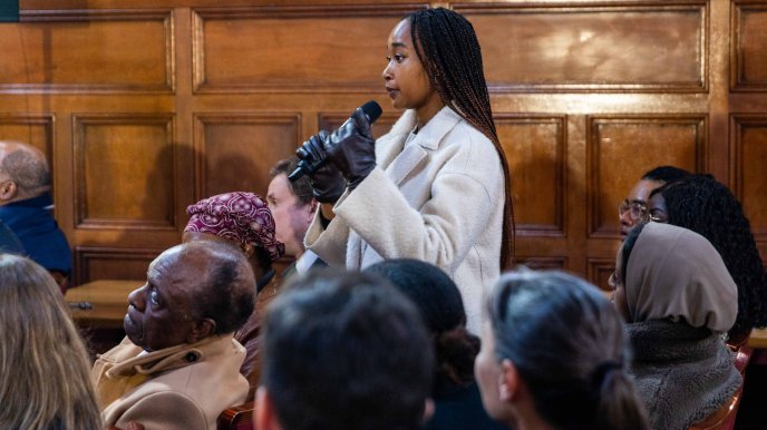 A young Black woman stands with a microphone to ask a question